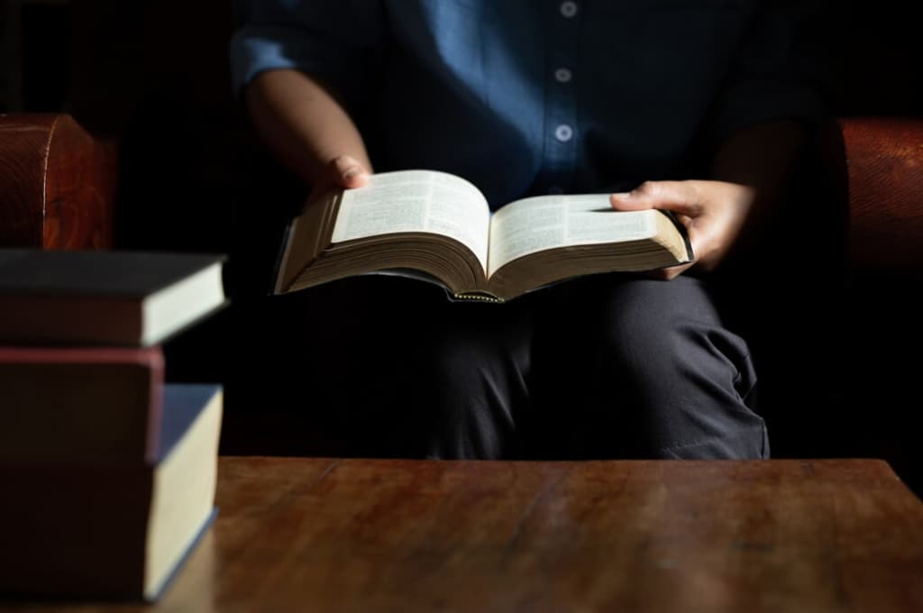 Person in einem blauen Hemd liest ein offenes Buch mit anderen Büchern daneben
